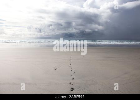 Frau im Abstand stehend gegen raue See auf sehr lange Strand unter dramatischen Wolkenhimmel mit geradem Anschluss von Abdrücken zu Ihr führen. Fossile Poi Stockfoto