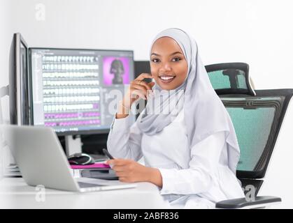 Junge afro-amerikanische moderne muslimische business Frau mit Kopftuch mit Kreativen hellen Büro Arbeitsplatz mit großen Bildschirm Stockfoto
