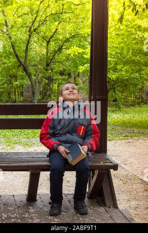 Träumen Happy Boy sucht, während allein sitzen auf Holzbank im Park mit Buch in der Hand. Selektive konzentrieren. Unfocused grünes Laub im Hinterg Stockfoto