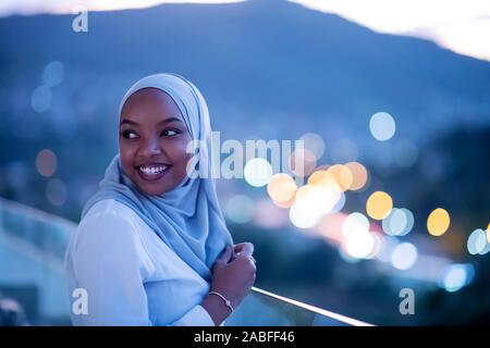 Afrikanische Moderne muslimische Frau in der Nacht am Balkon lächelnd an Kamera mit Stadt bokeh Lichter im Hintergrund Stockfoto