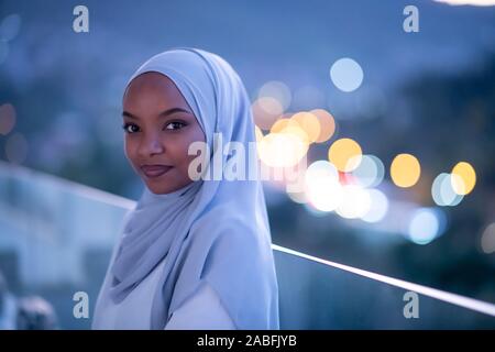Afrikanische Moderne muslimische Frau in der Nacht am Balkon lächelnd an Kamera mit Stadt bokeh Lichter im Hintergrund Stockfoto