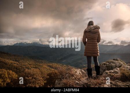 Eine Frau in einem braunen Mantel mit Pelzkragen stehen auf einem Hügel in Korsika und genießen den Blick auf die schneebedeckten Berge in der Ferne Stockfoto