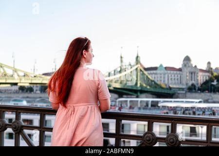 Ein rothaariges Mädchen Tourist in einem zarten rosa Kleid steht durch den Fluss und schaut auf die Eiserne Brücke bei Sonnenuntergang. Stockfoto