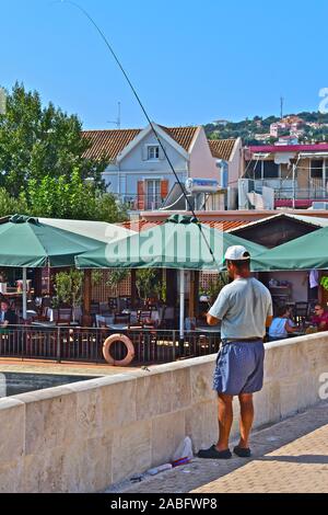 Ein Mann angeln in der De Bosset Brücke, ein Stein causeway Verknüpfung von Argostoli mit Drapano, auf der anderen Seite der Bucht. Waterside Cafe hinter sich. Stockfoto