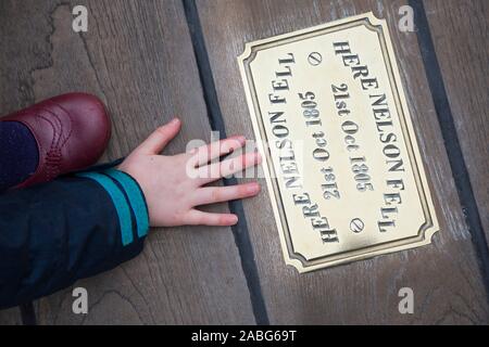 Kind Gast/kid Touristische untersuchen die Metall Messingschild auf der Website von HMS Victory an der Stelle, wo Admiral Lord Nelson fiel nach geschossen wird von einem französischen Musketier. Portsmouth. Großbritannien (105) Stockfoto