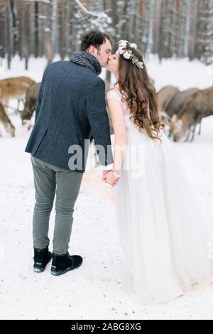 Jungen lieben schöne Hochzeit paar Küssen und halten sich an den Händen, Outdoor im Winter in einem stilvollen Hochzeit Kleidung. Hirsch auf dem Hintergrund. Schönheit Stockfoto