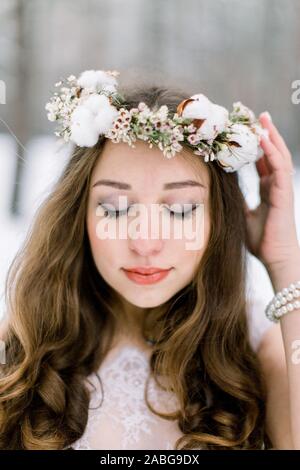 Schöne Braut mit lockigem Haar, ihr Blumen Kopf band Kranz berühren, von Blumen und Baumwolle, stehen im Freien, auf dem Hintergrund der Winter Stockfoto