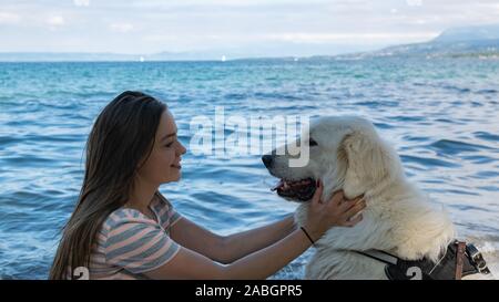 Lächelnde junge Frau mit hellbraunem Haar hält einen großen, weißen Hund auf einen unscharfen Hintergrund der Blaue See und die Berge. Stockfoto