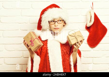 Junge süße santa claus Junge mit Brille im roten Pullover und das neue Jahr hat mit dekorativen Weihnachts- oder Weihnachten Strumpf oder Boot holding Geschenke in Handwerk Papier auf weißen Mauer Hintergrund gewickelt. Stockfoto