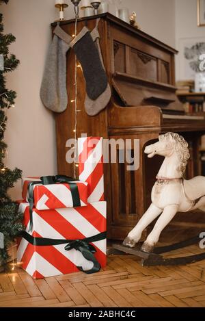 Bunte Weihnachtsgeschenke verpackt in einem warmen Braun Vintage Retro Interieur. Geschenke auf dem Hintergrund der Piano, ein Baby Schaukelpferd und Weihnachten s Stockfoto