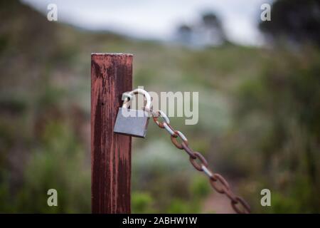 Alte und rostige Kette mit einem geschlossenen Vorhängeschloss im Feld mit grünen Bäumen Hintergrund Stockfoto