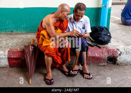 Ein buddhistischer Mönch und lokaler Mann Plaudern auf der Plattform eines Bahnhof, Yangon, Myanmar. Stockfoto