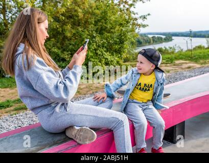 Junge Mutter nimmt Bilder der Boy ist ihr Sohn, in seiner Hand ein Handy, Emotionen, Freude, Spaß und Entspannung am Wochenende. Wenig junge spielt Skateboard Stockfoto