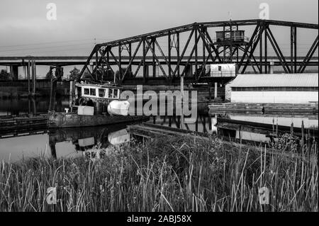 Alten, verlassenen Tug Boat in industriellen Marina mit train trestle im Hintergrund in Schwarz und Weiß. Stockfoto