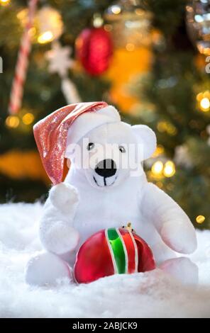 Eine kleine weiße Eisbär trägt eine rote Weihnachtsmütze und hält eine glänzende rote und grüne Verzierung Stockfoto