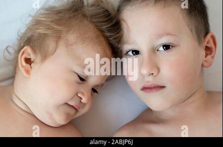 Close-up Portrait von Geschwister an einander suchen und liegend auf dem Bett. Gesunde junge liebt es, seine Gäste mit kleinen Bruder. Zerebralparese Baby mit Bruder Stockfoto