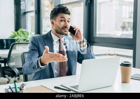Positive Geschäftsmann zeigte mit Hand am Laptop während des Sprechens auf dem Smartphone Stockfoto