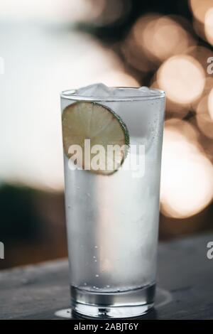 Cocktail Glas mit Gin Tonic und ein Kalk- und Eiswürfel auf einem Balkon mit malerischem und herrlichen Blick auf die Küste des Mittelmeers bei Sonnenuntergang i Stockfoto