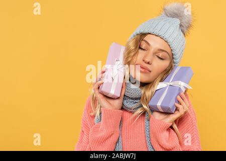 Frau mit Hut und Schal mit geschlossenen Augen holding Geschenkboxen isoliert auf Gelb Stockfoto