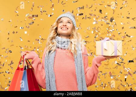 Glückliche Frau in hat Holding Geschenkbox und Einkaufstaschen unter fallenden Konfetti isoliert auf Gelb Stockfoto