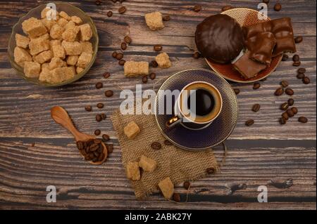 Eine Tasse Kaffee, Kaffeebohnen, braunen Zucker in einer Schüssel Zucker, Schokolade Stücke, Marshmallows in Schokolade auf einem hölzernen Hintergrund Stockfoto
