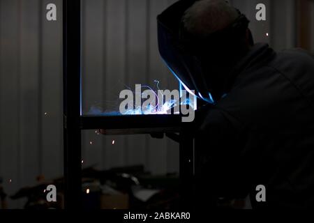 Fokussierte Schweißer Designer das Tragen eines Helmes und wokring an einem Projekt durch Schweißen Teile aus Metall, während Sie schöne Rauch in seiner Werkstatt in der Nacht Stockfoto