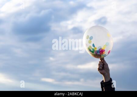 Eine Hand mit einem durchsichtigen Ballon mit viele bunte Konfetti auf der Innenseite mit blauem Himmel und Wolken im Hintergrund horizontal Stockfoto