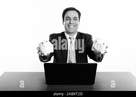 Studio shot der Jungen glücklich Persischen Geschäftsmann lächelnd, während sie zwei Spardosen mit Laptop auf hölzernen Tisch Stockfoto