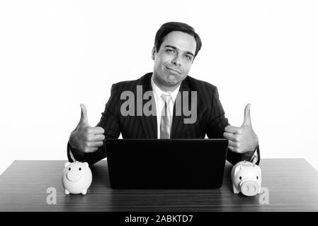 Studio shot der jungen Persischen Geschäftsmann Daumen hoch geben mit Laptop und zwei spardosen auf hölzernen Tisch Stockfoto