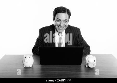 Studio shot der Jungen glücklich Persischen Geschäftsmann Lächeln beim Sitzen mit Laptop und zwei spardosen auf hölzernen Tisch Stockfoto