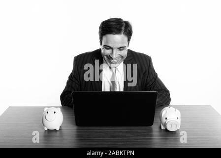 Studio shot der Jungen glücklich Persischen Geschäftsmann lächelnd, während sie Laptop mit zwei spardosen auf hölzernen Tisch Stockfoto