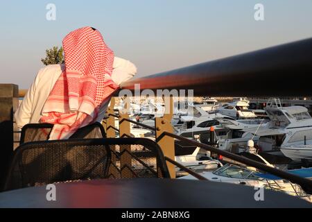 Kuwaitischen Mann mit roten und weißen Kopf Kleid (palituch) Hinter sitzen mit Blick auf eine Marina mit Boote gesehen Stockfoto