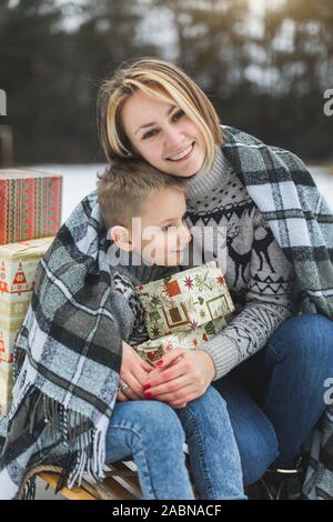 Mutter und Sohn genießen schneereiche Winter Tag im Freien sitzen auf dem Schlitten, umarmen, mit Plaid und Holding Weihnachtsgeschenke abgedeckt Stockfoto