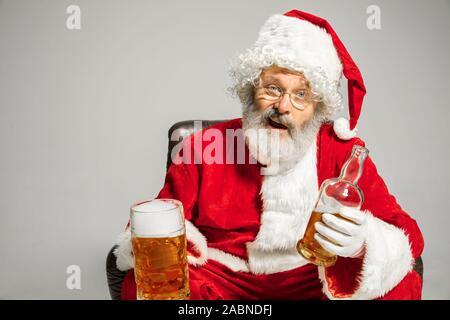 Santa Claus Bier sitzen auf Sessel, Glückwunsch, sieht betrunken und glücklich. Kaukasische männliches Modell in der traditionellen Tracht. Neues Jahr 2020, Geschenke, Urlaub, Winter Stimmung. Copyspace für Ihre Anzeige. Stockfoto