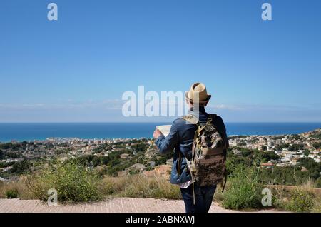 Ein junger Mann mit Rucksack und eine Karte der Gegend in seinen Händen steht auf einem Berg in der Nähe von Stadt und Meer, Rückansicht Stockfoto