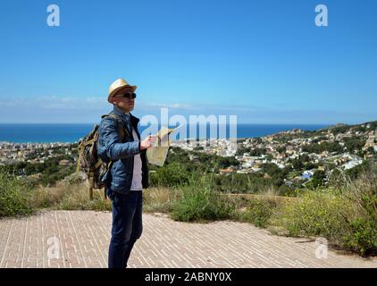 Ein Tourist mit einem Rucksack und eine Karte der Gegend in seinen Händen steht auf einem Berg in der Nähe der Stadt und das Meer. Stockfoto