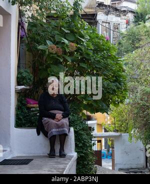 In Pogradec, Albanien - September 26, 2019: ältere Frau trägt einen Rock, schwarze Bluse, schwarze Strickjacke sitzt auf einem niedrigen weißen Wal Stockfoto