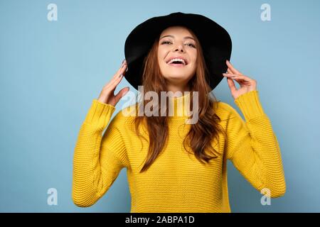 Eine Brünette in einem gelben Pullover steht vor einem blauen Hintergrund, lacht mit ihren Kopf nach hinten geworfen und hält den Hut mit ihren Händen Stockfoto