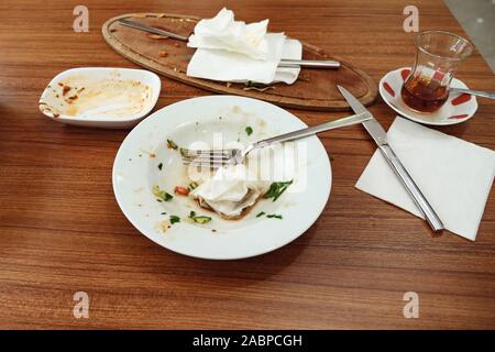 Leere schmutzigen weißen Platte oder Teller, Tasse Tee, Löffel, Messer, Gabel, und Abfall essen nach auf braune Holztisch Hintergrund gegessen Stockfoto