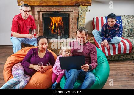 Große Familie verbringen die Zeit zusammen in der Nähe von Kamin: Erwachsene sind, Wein trinken und auf den Bildschirm des Notebooks beim Sitzen auf dem Sitzsack, Stockfoto