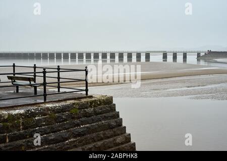 Teilweise mit Blick auf die aus Stein gebaute Pier und Sitz mit einem Eisenbahnviadukt Überqueren einer Flussmündung im Hintergrund auf einem nassen und trüben Tag Stockfoto