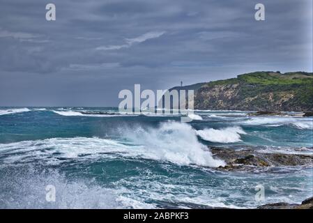 Landschaft Foto von rauer See mit Wellen im Vordergrund und grünen Klippen im Hintergrund mit einem hellen Haus auf. Stockfoto