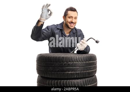 Fröhliche Automechaniker Gestik großartige mit Hand und hält Schraubenschlüssel hinter dem Auto Reifen auf weißem Hintergrund Stockfoto