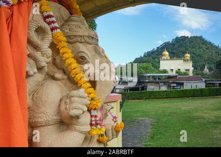 Eine Statue von hinduistischen Elefantengott Ganesh an Thendayuthapani (Hindu) Tempel in der Stadt Phuket, Thailand; b/g: Gurdwara Siri Guru Singh Sabha (Sikh Tempel) Stockfoto