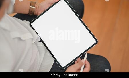 7/8 Schuß des jungen Mannes mit seinem leeren Bildschirm digital tablet, während im komfortablen Zimmer sitzen Stockfoto