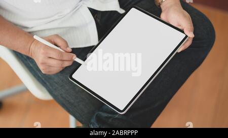 7/8 Schuß des jungen Mannes mit seinem leeren Bildschirm digital tablet, während im komfortablen Zimmer sitzen Stockfoto