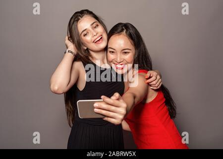 Junge Frauen tragen Abendkleid studio Ständigen umarmen auf grauem Hintergrund holding Smartphone unter selfie Foto Kamera posieren isoliert lächelnd Stockfoto