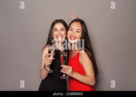 Junge Frauen tragen Abendkleider Studio stehen auf grauen Hintergrund halten Gläser Champagner posiert für die Kamera angezeigt Gruß Geste l isoliert Stockfoto