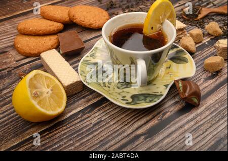 Eine Tasse schwarzer Tee, in Scheiben geschnittene Zitronen, brauner Zucker, Scheiben, Haferflocken Kekse, Waffeln auf einem hölzernen Hintergrund. Nahaufnahme Stockfoto