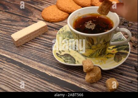 Die Hand des Mädchens legt ein Stück braunen Zucker in einer Tasse schwarzen Tee, oatmeal Cookies, Tee, Waffeln, brauner Zucker auf einer hölzernen Hintergrund. Nahaufnahme Stockfoto